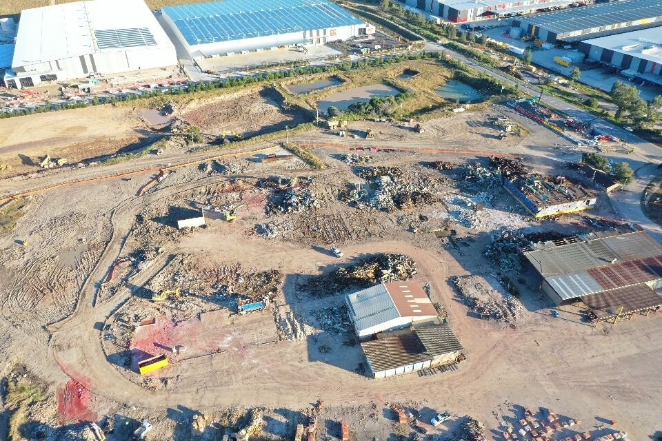 An aerial view of an industrial site reveals scattered piles of debris, punctuated by bricks and pavers. A few small buildings, roads, and warehouses are neatly arranged nearby.