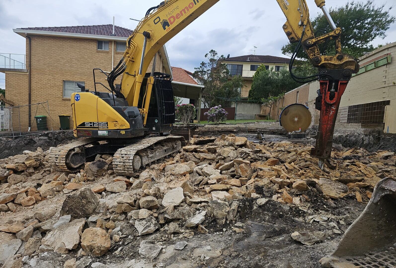 A yellow excavator with a large drill attachment breaks up rocks at the bustling construction site on 63 Johnston Parade, surrounded by residential buildings.