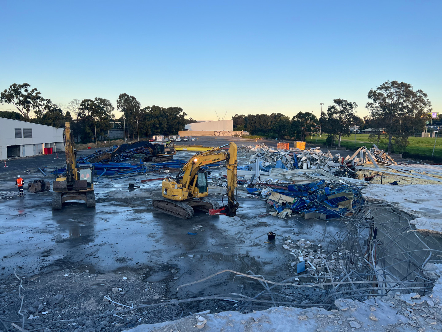 A construction site featuring two excavators methodically providing demolition services amid the rubble and debris, surrounded by trees and a building, all under a clear blue sky.