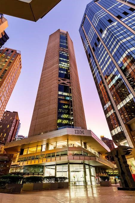 Tall skyscraper in an urban setting with reflective glass buildings surrounding it and an illuminated sky in the background.