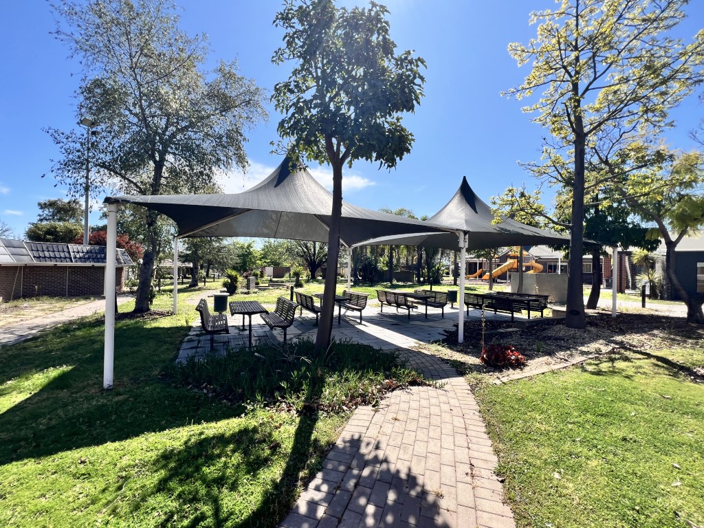 Outdoor seating area with shade sails, surrounded by trees and greenery on a sunny day. Brick pathway leads to tables and chairs. Playground equipment in the background.